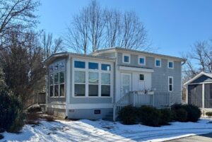 Charming small gray house on Drifting Brook Lane, with many windows, porch, and light snow under a clear sky.