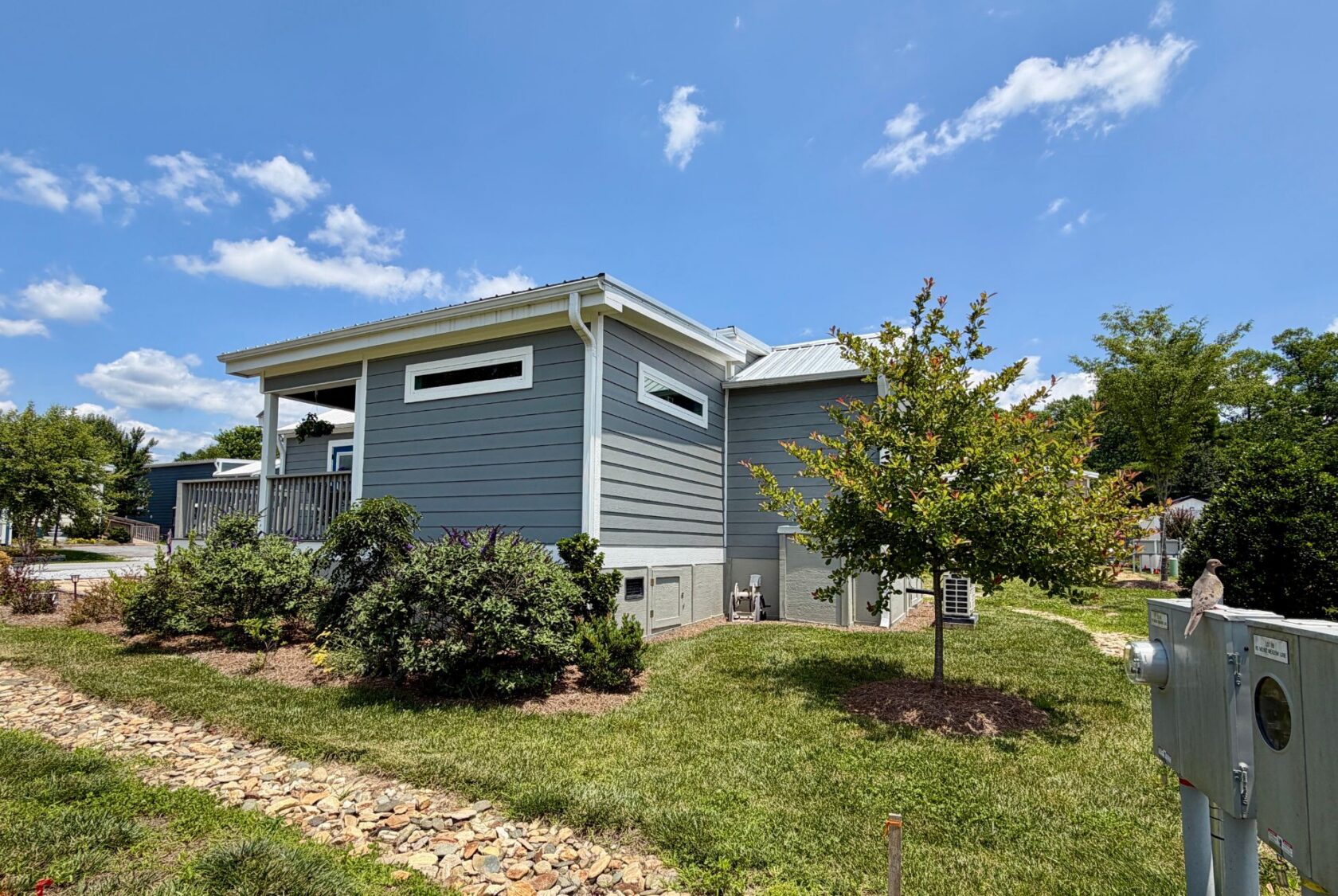 A modern gray house on Mount Meadow Lane with a small porch, green grass, trees, and shrubs under a blue sky.