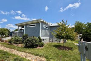 A modern gray house on Mount Meadow Lane with a small porch, green grass, trees, and shrubs under a blue sky.
