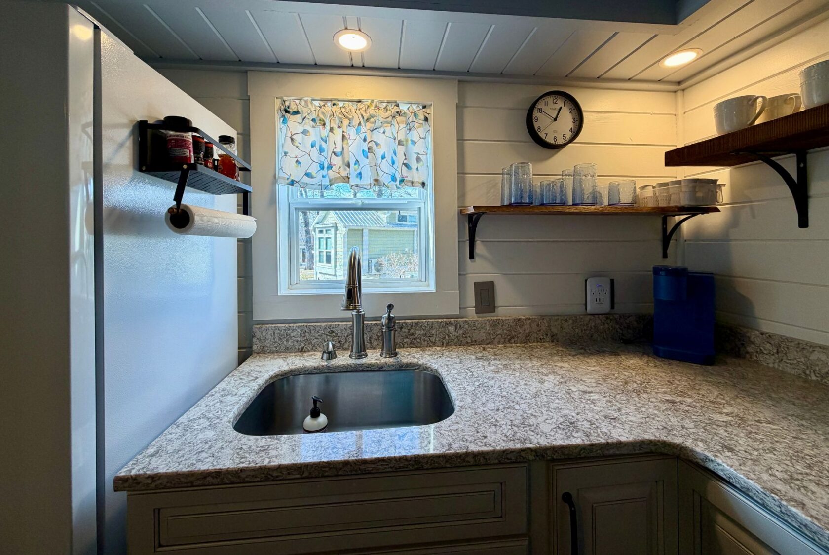 A cozy Empire Lane kitchen corner with a sink, countertop, shelves, and a window with a floral curtain.