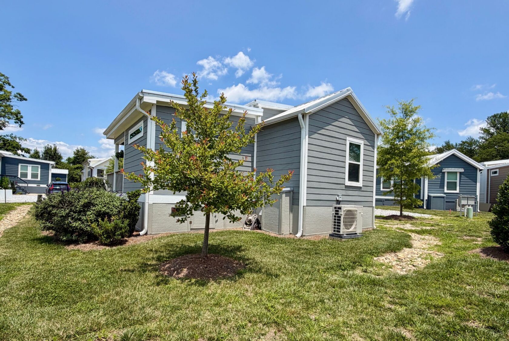 A small gray house with white trim and a young tree sits on Mount Meadow Lane under a blue, cloud-streaked sky.
