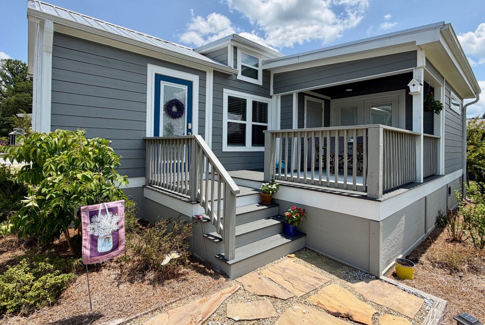 Gray house on Mount Meadow Lane with a small porch, blue door, potted plants, and a decorative flag in the yard.
