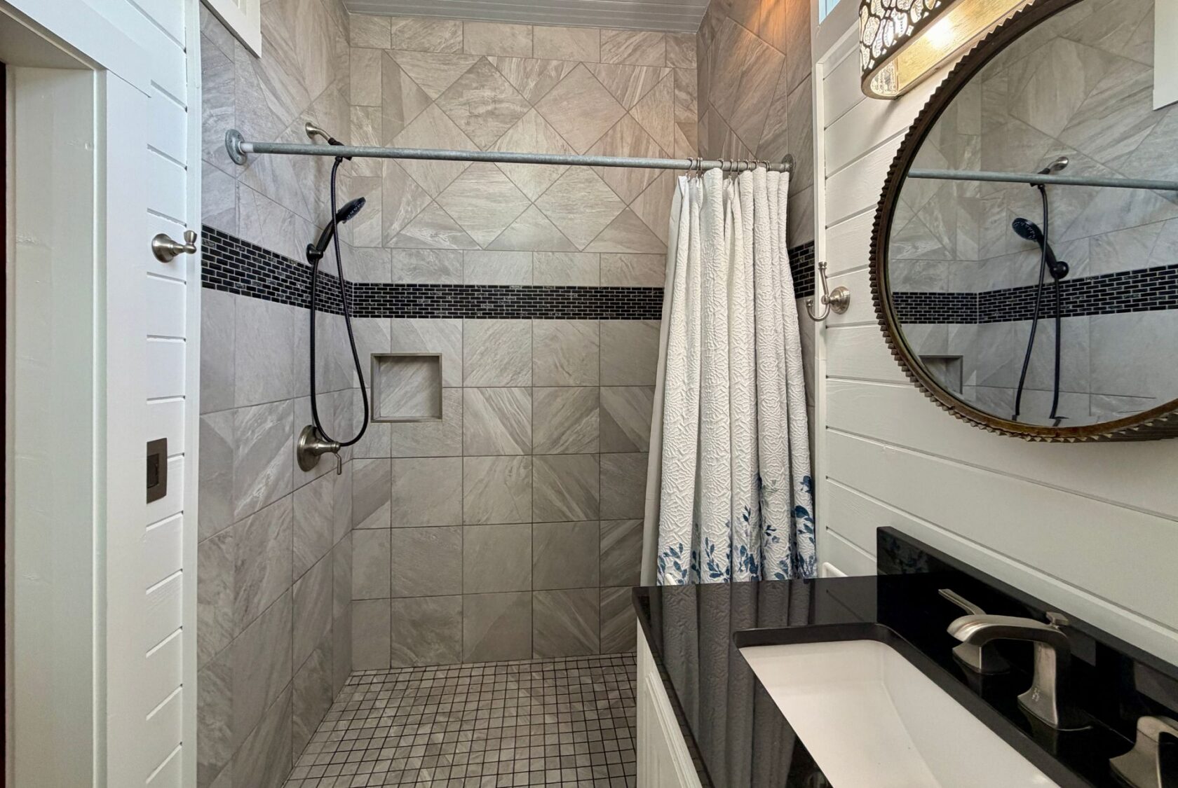 Modern bathroom on Empire Lane with a tiled walk-in shower, patterned curtain, and a round mirror above a black countertop.