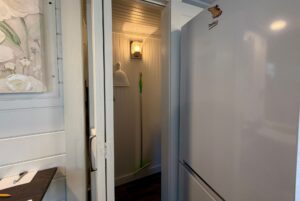 A small closet with a light at 5 Empire Lane, holding a broom and dustpan, beside a refrigerator and counter.