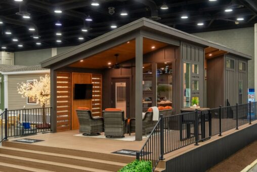 Modern tiny home display with a covered porch, outdoor seating, and railing at an indoor exhibition.