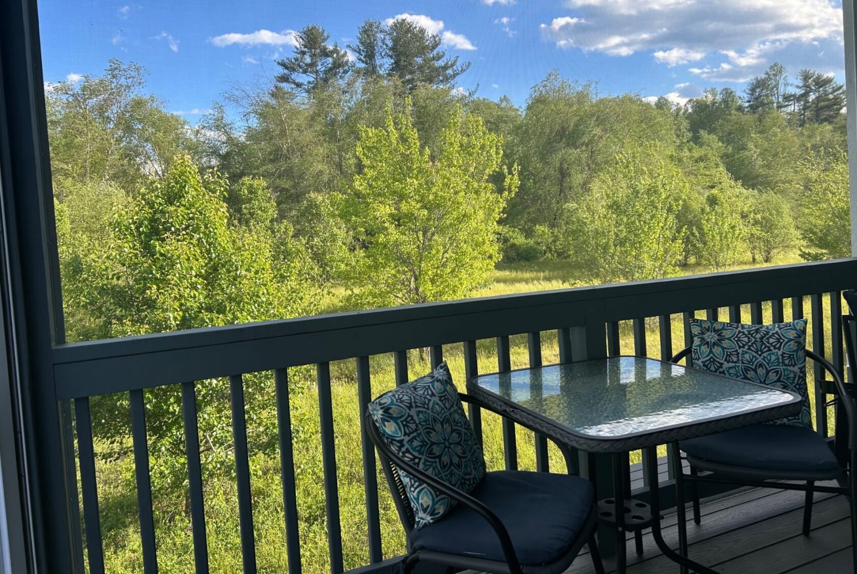 A glass table with two chairs on a porch at Mount Meadow Lane overlooks a lush green landscape under a blue sky.