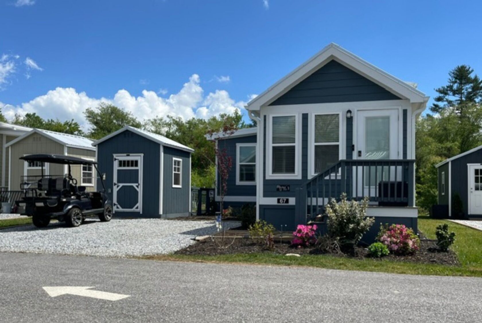 Blue tiny house on Mount Meadow Lane with porch, garden, shed, and golf cart on a gravel driveway under a blue sky.