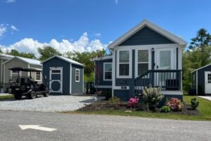 Blue tiny house on Mount Meadow Lane with porch, garden, shed, and golf cart on a gravel driveway under a blue sky.
