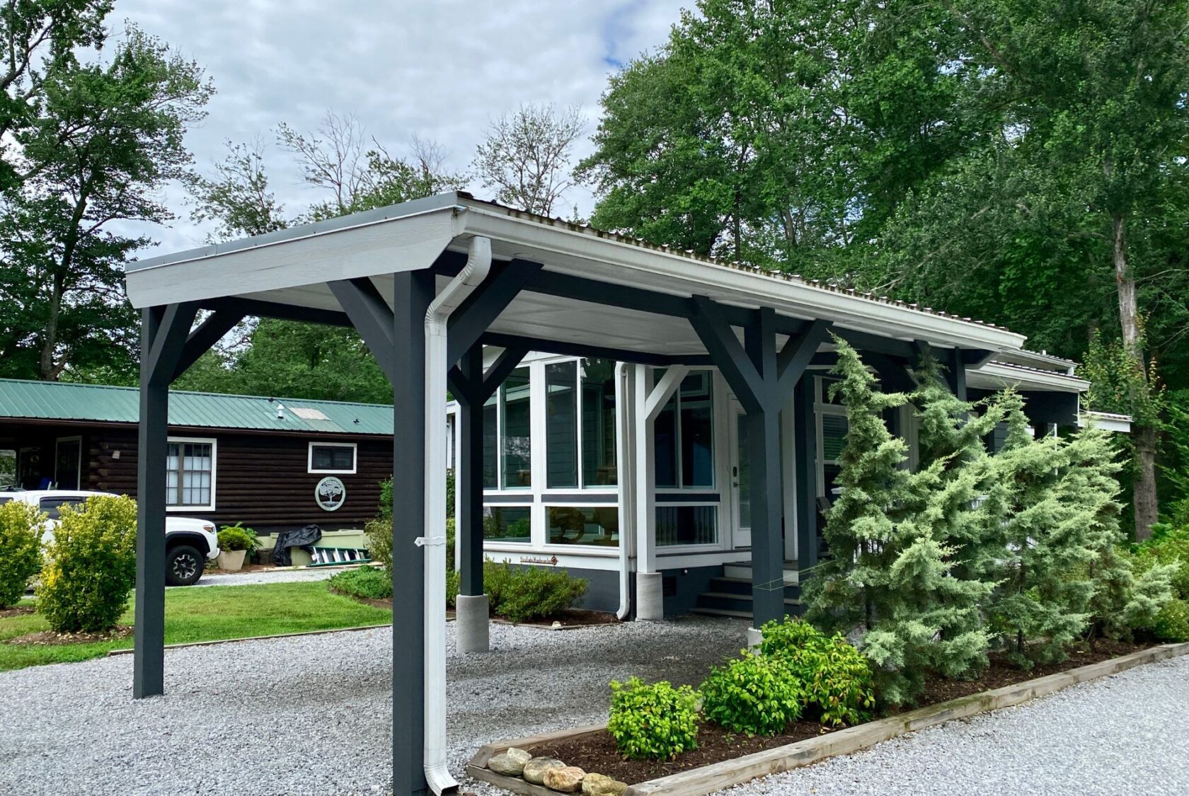 A modern gray carport on Just Wandering Lane with a gravel driveway, beside a small house and lush trees.
