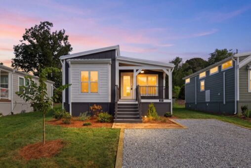 Small modern house with front porch, lit windows, gravel driveway, and landscaped yard at sunset.
