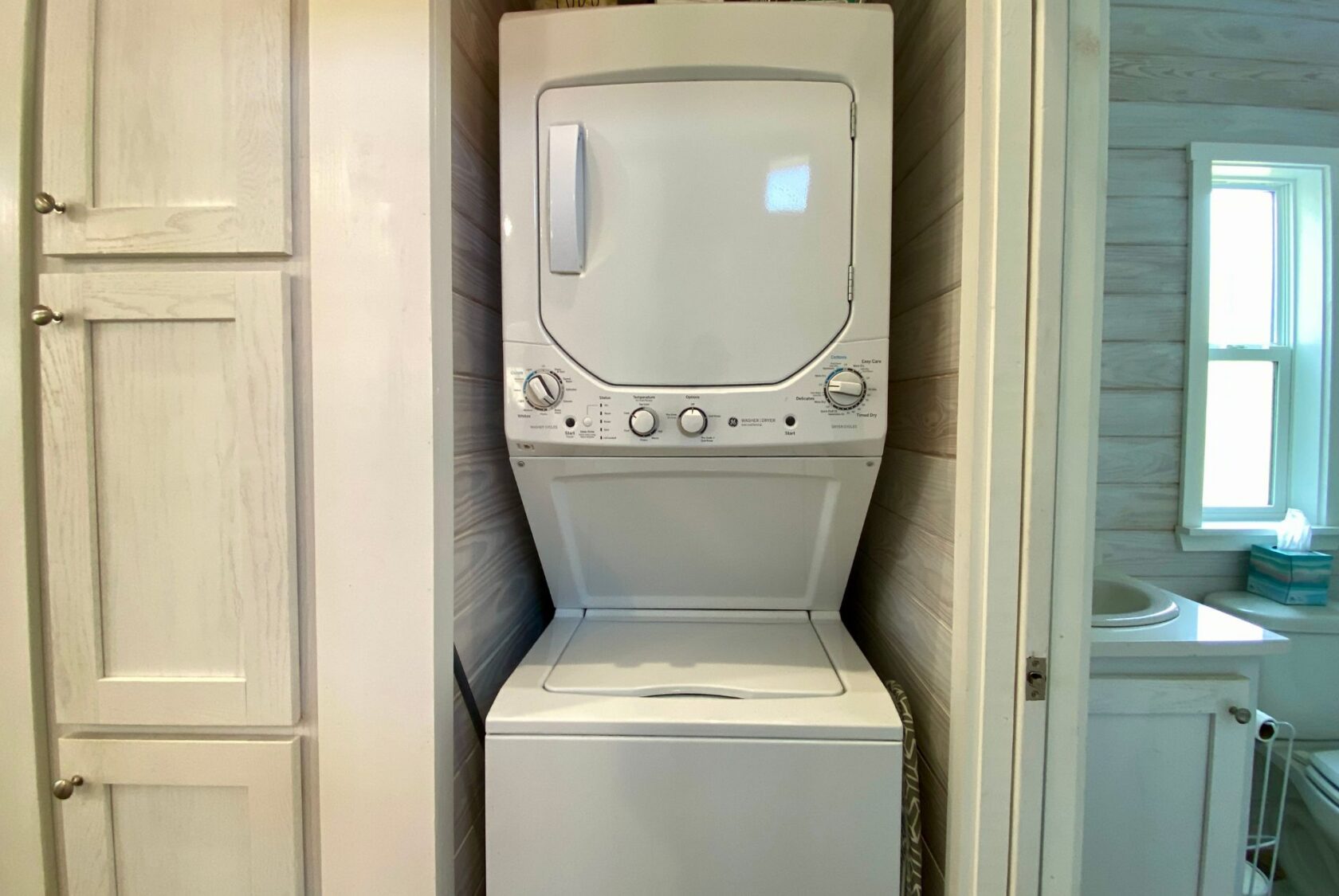 Stacked washer and dryer unit on Easy Lane in a small laundry nook next to a bathroom with a window and sink.