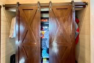 Two partially open barn doors at 31 Rest Awhile Lane reveal a cluttered closet filled with clothes and linens.