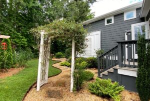 Wooden garden arbor with climbing plants leads to Just Wandering Lane, a curved gravel path by a gray house with white trim.