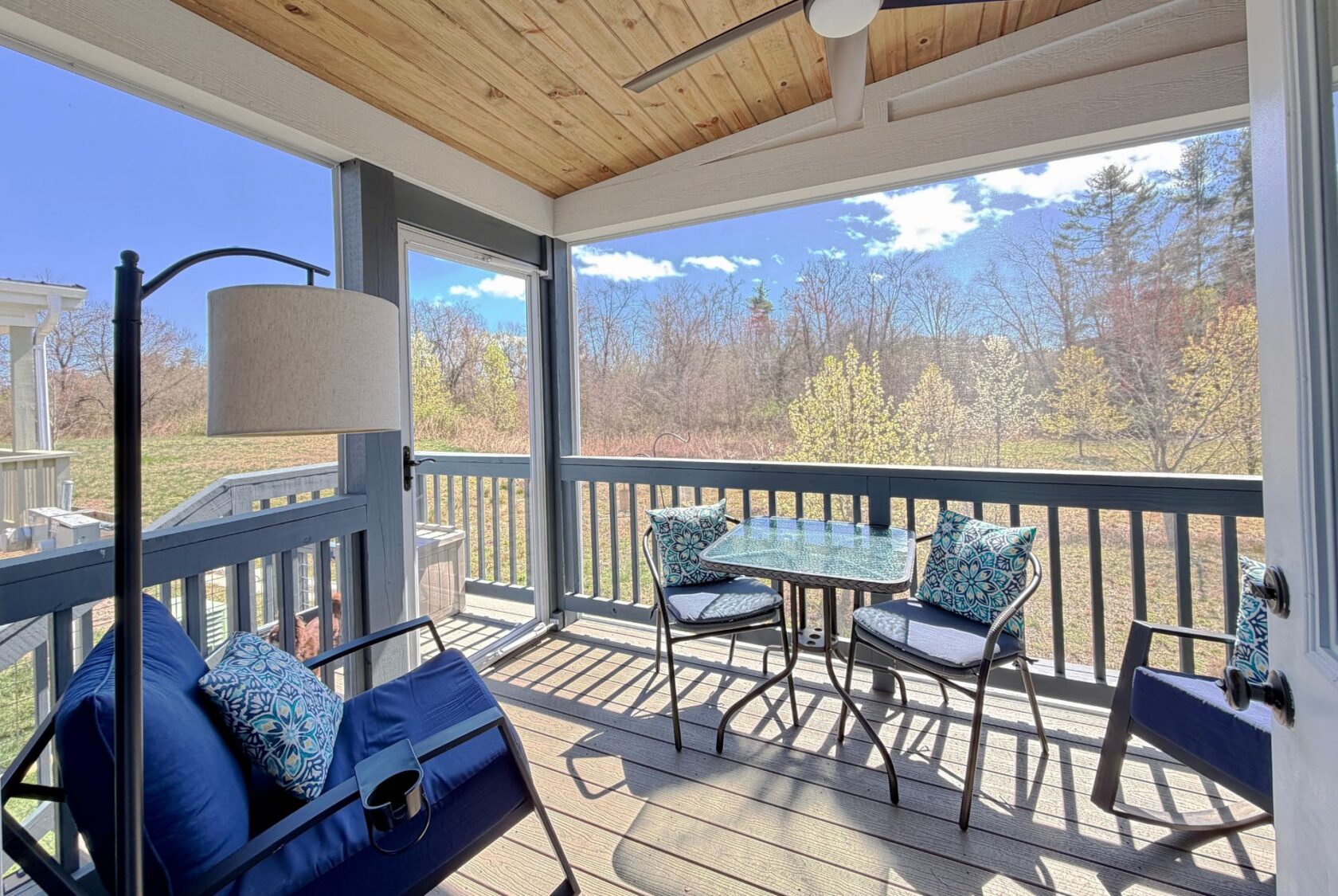 A cozy screened porch on Mount Meadow Lane with a cushioned bench, table, and chairs overlooking a sunny backyard with trees.