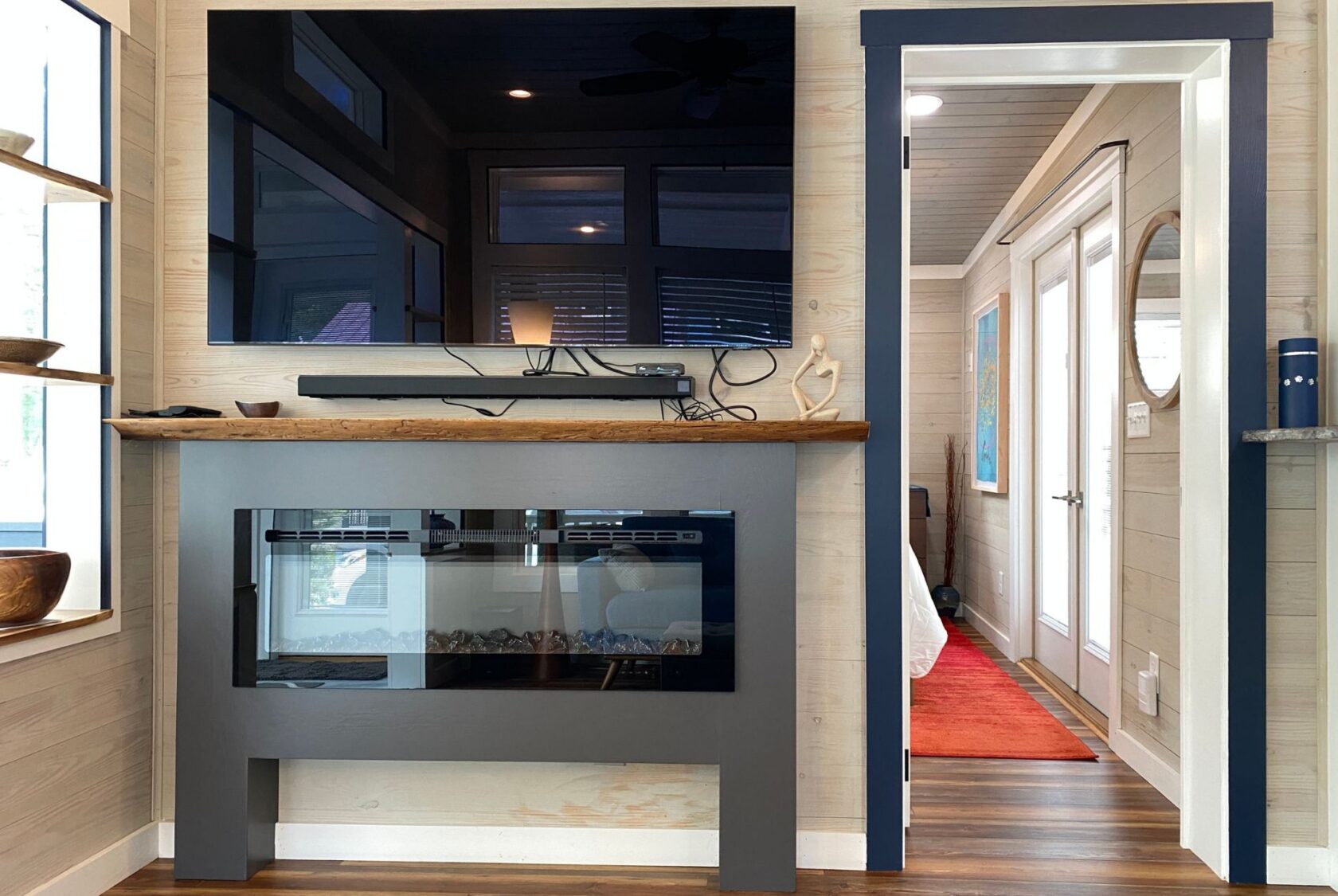 A modern living room on Just Wandering Lane features a wall-mounted TV above an electric fireplace and a bright bedroom doorway.