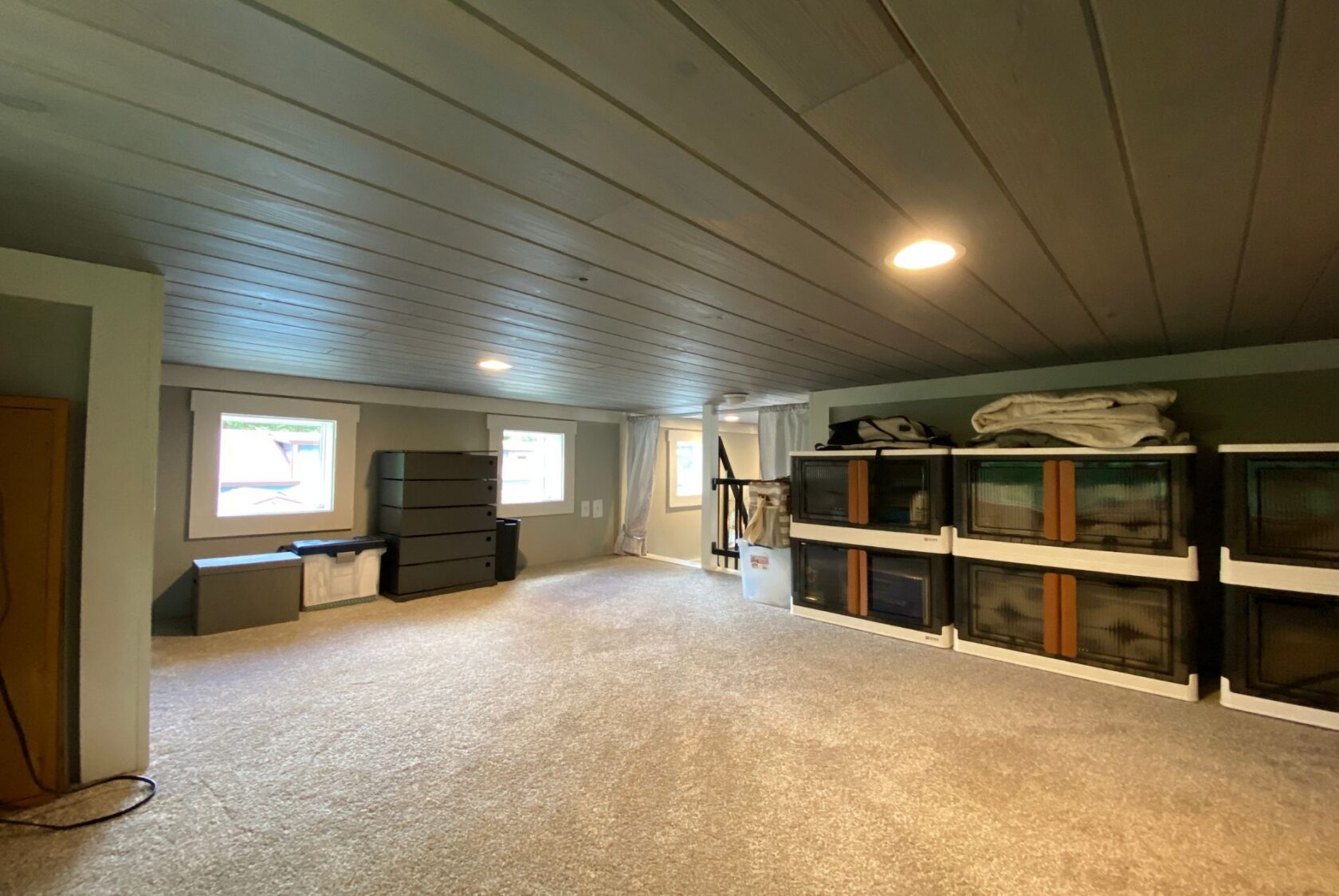 Spacious attic room with carpet, storage bins, and two windows on Just Wandering Lane; folded blankets on shelves.