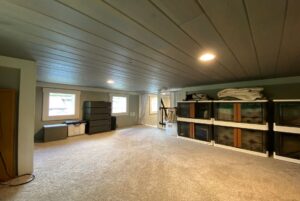 Spacious attic room with carpet, storage bins, and two windows on Just Wandering Lane; folded blankets on shelves.