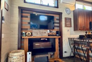 A wall-mounted TV above an electric fireplace on Rest Awhile Lane, with rustic decor and kitchen area to the right.
