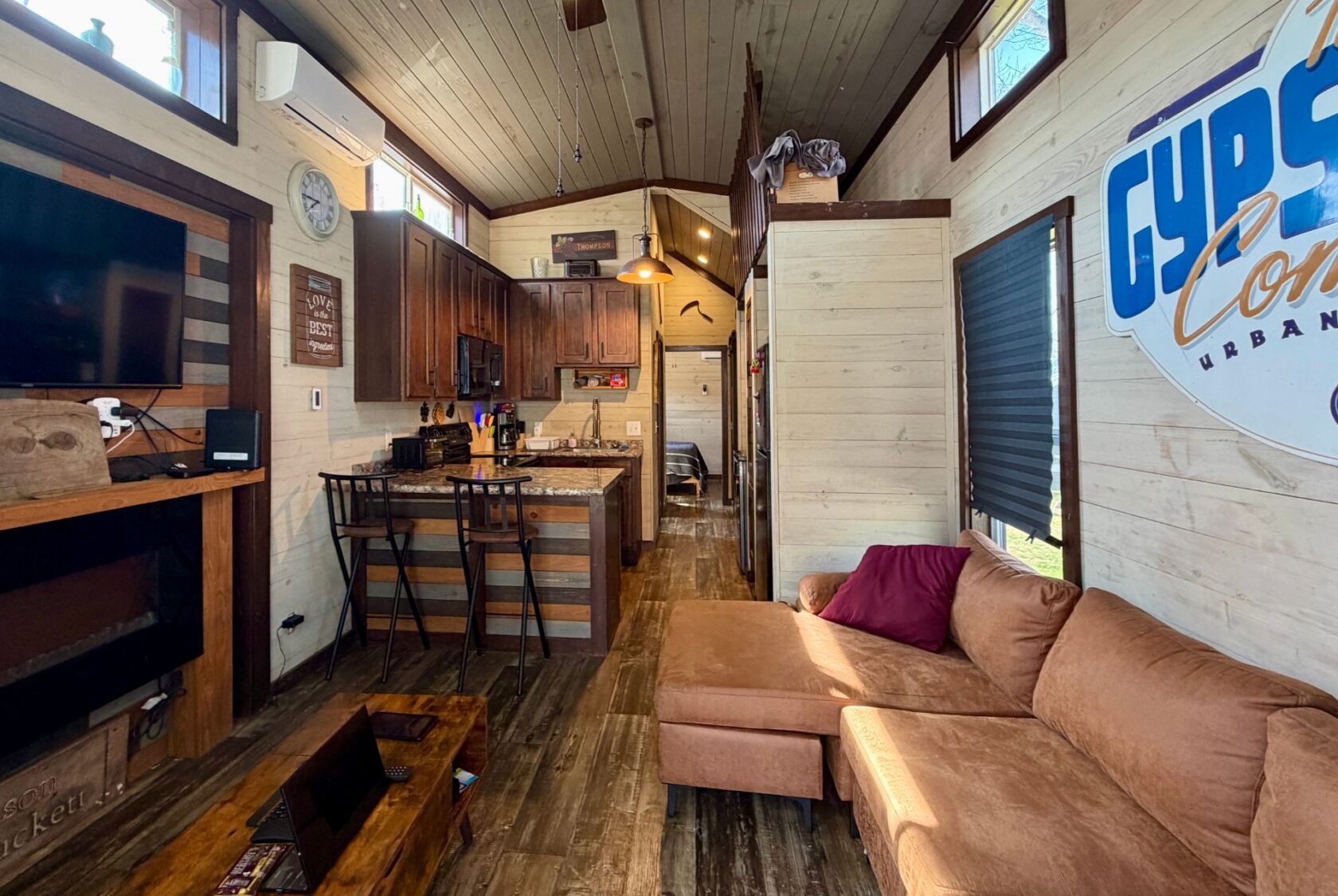 Cozy tiny house interior at 31 Rest Awhile Lane with kitchen, wooden floors, brown sofa, and sunlight through the windows.