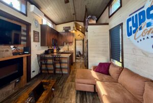 Cozy tiny house interior at 31 Rest Awhile Lane with kitchen, wooden floors, brown sofa, and sunlight through the windows.