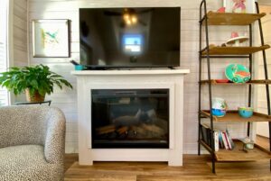 A TV on a white fireplace with a plant, shelf, and books sets an Easy Lane vibe in this cozy living room.