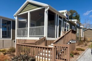 Green tiny house on Breezy Meadow Lane with a screened porch, brown railing, and steps under a clear blue sky.