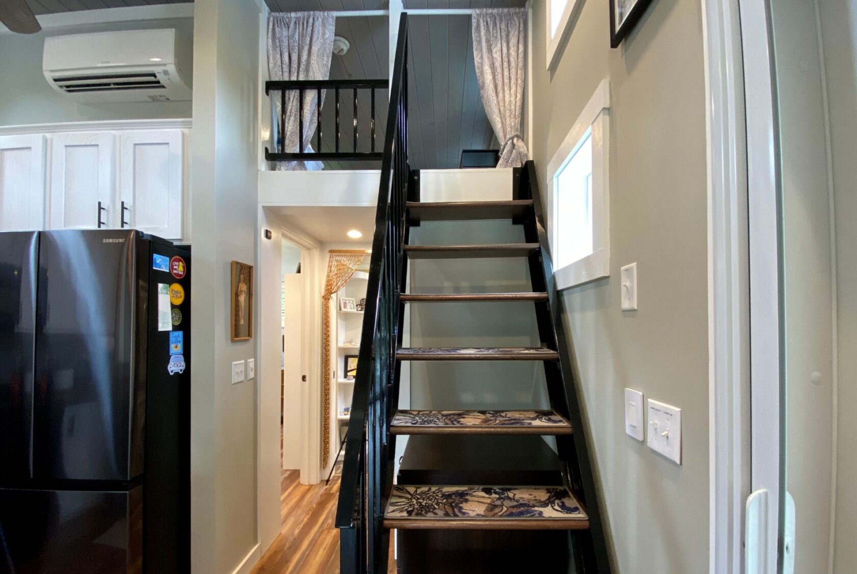Modern tiny house interior on Just Wandering Lane with black staircase, loft, kitchen appliances, and wooden flooring.