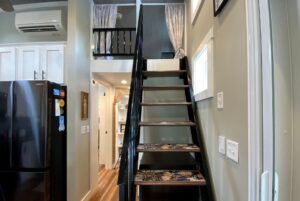 Modern tiny house interior on Just Wandering Lane with black staircase, loft, kitchen appliances, and wooden flooring.