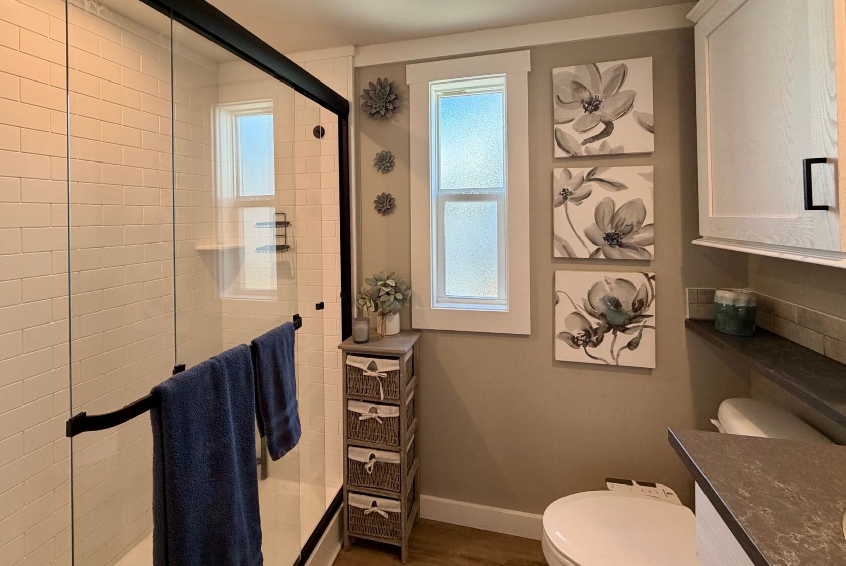 Modern bathroom on Mount Meadow Lane with glass shower, blue towels, floral wall art, and wicker storage by a frosted window.