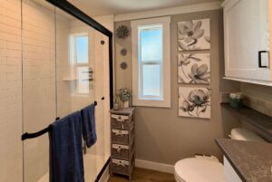 Modern bathroom on Mount Meadow Lane with glass shower, blue towels, floral wall art, and wicker storage by a frosted window.