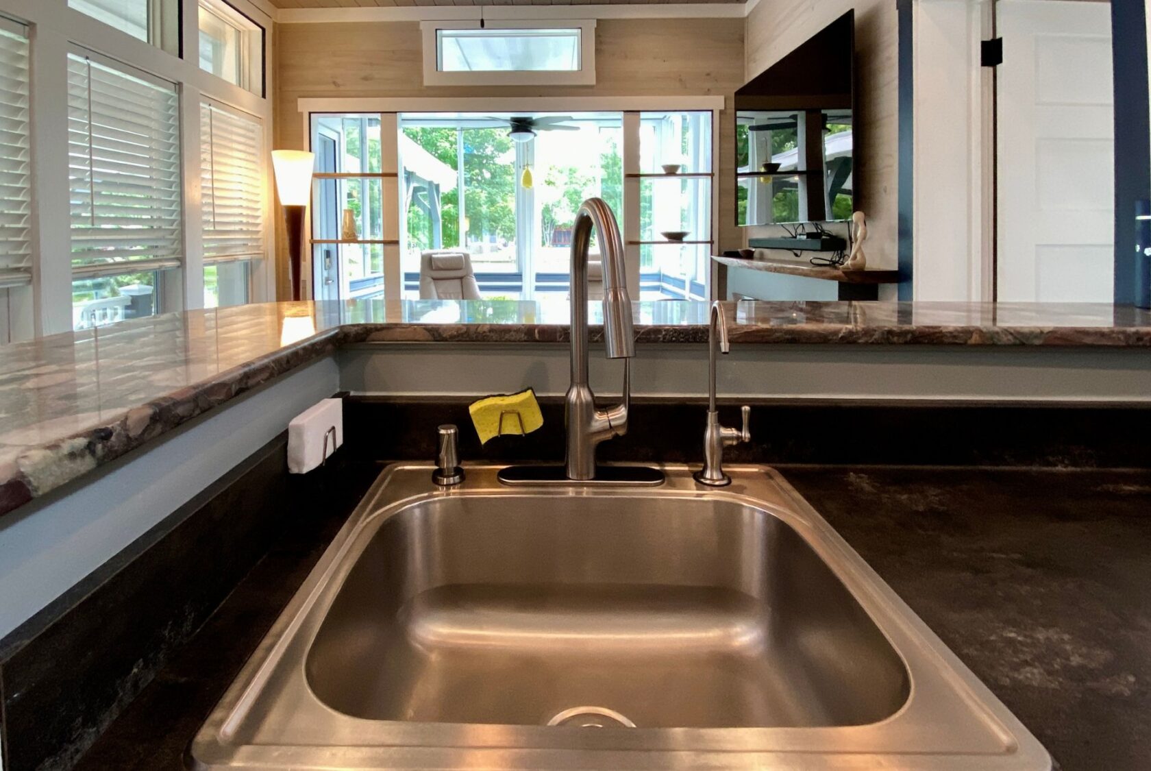 Close-up of a kitchen sink with a modern faucet at Just Wandering Lane, overlooking a bright living area with large windows.
