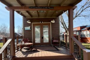 A small wooden house at 31 Rest Awhile Lane features a porch, chairs, and a sign that reads Love Grows Best in Little Houses.