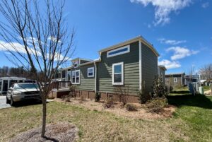 Green tiny house with white trim on Breezy Meadow Lane, bare trees, and blue sky in a suburban neighborhood.