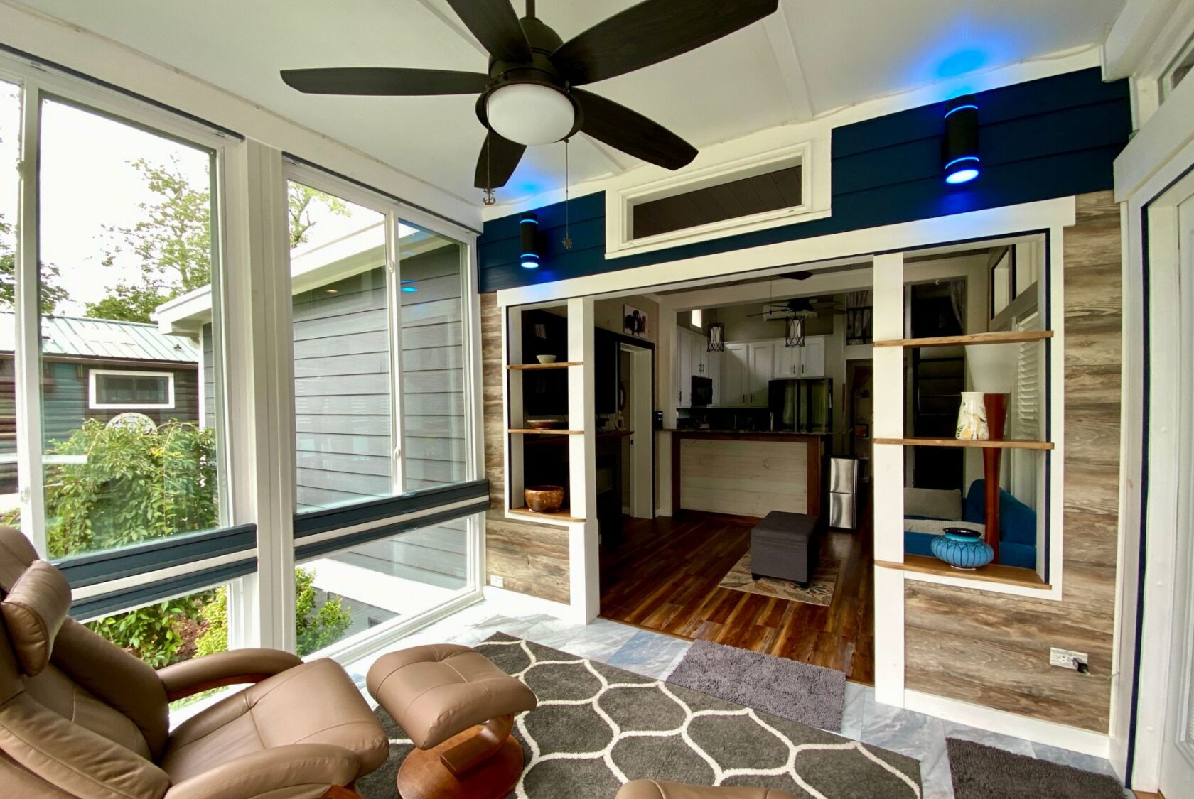 Modern sunroom on Just Wandering Lane with recliners, ceiling fan, large windows, and a view into the cozy kitchen and living area.