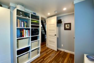 White shelving with books and baskets in a blue room on Just Wandering Lane, featuring wood floors and an open door.