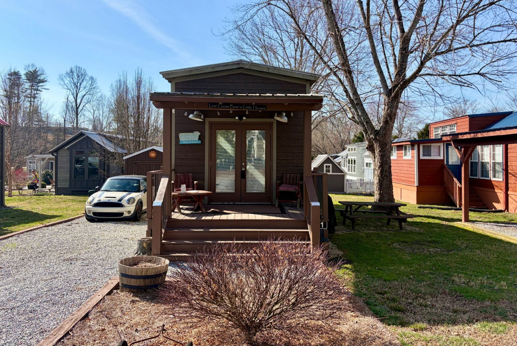 A tiny house with a front porch sits on Rest Awhile Lane, near a parked car, surrounded by trees and small homes.