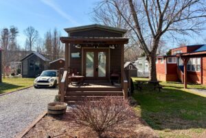 A tiny house with a front porch sits on Rest Awhile Lane, near a parked car, surrounded by trees and small homes.