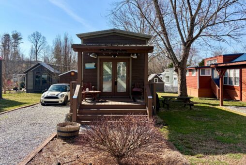 A tiny house with a front porch sits on Rest Awhile Lane, near a parked car, surrounded by trees and small homes.