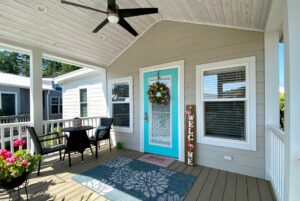 A cozy porch on Easy Lane with a turquoise door, welcome sign, flowers, table, chairs, and ceiling fan.
