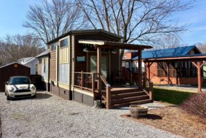 A small brown tiny house with a porch at 31 Rest Awhile Lane, next to a Mini Cooper on a gravel driveway.