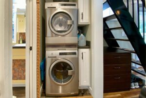 Stacked washer and dryer in a small laundry nook beside Just Wandering Lane's dark staircase and white cabinets.