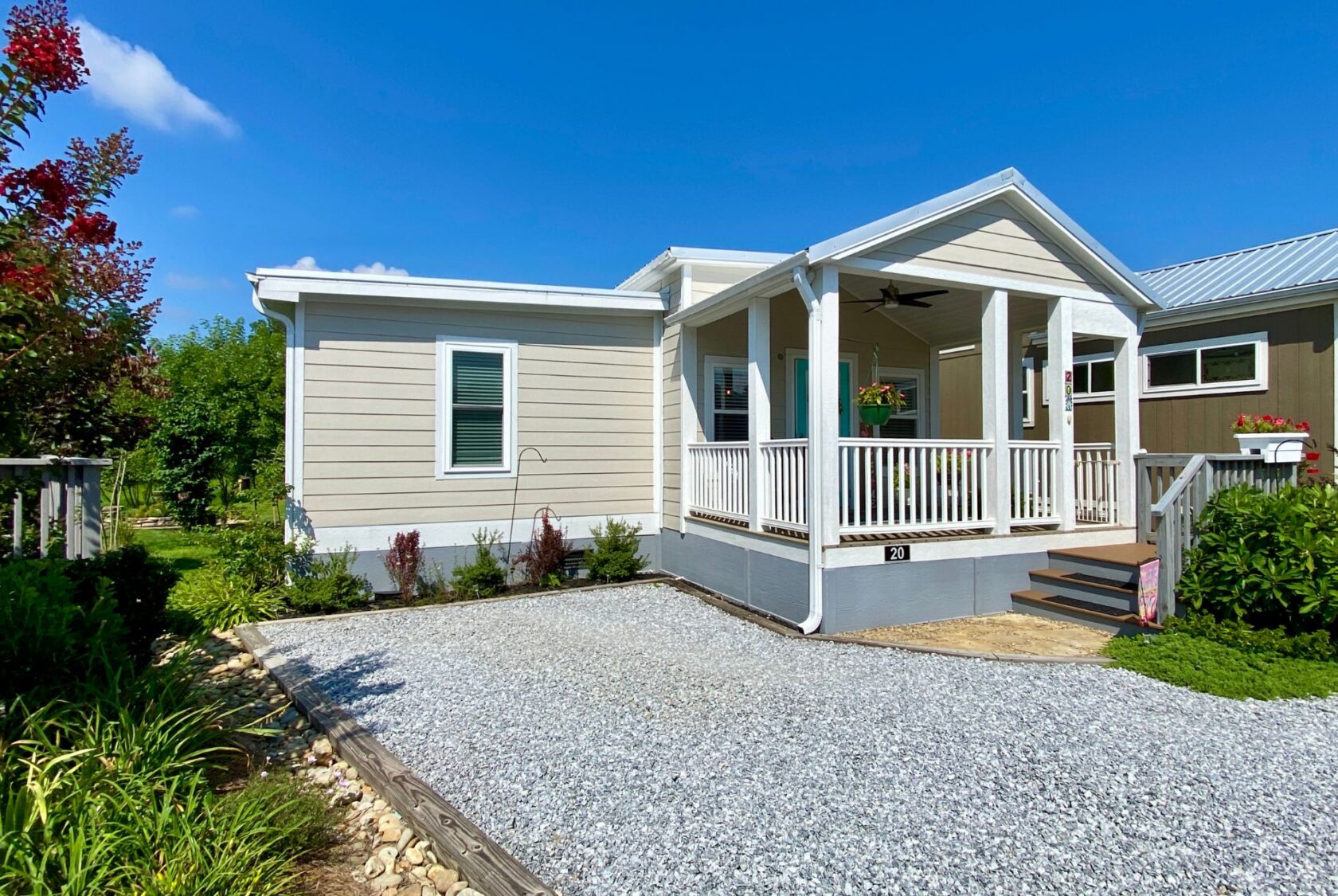 Small beige house on Strolling Easy Lane with a white porch, gravel driveway, and landscaping under a bright blue sky.