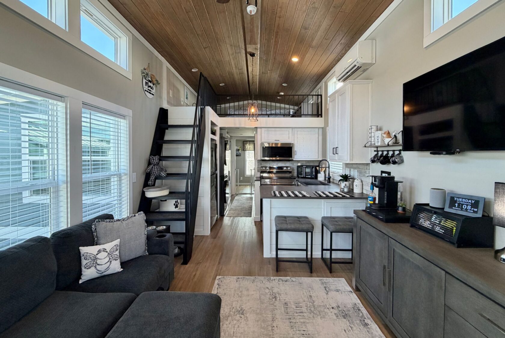Modern tiny house interior on Mount Meadow Lane with loft, kitchen, gray sofa, TV, and large windows letting in natural light.