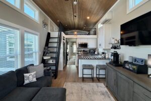 Modern tiny house interior on Mount Meadow Lane with loft, kitchen, gray sofa, TV, and large windows letting in natural light.
