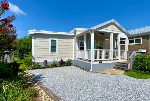 Small beige house on Strolling Easy Lane with a white porch, gravel driveway, and landscaping under a bright blue sky.