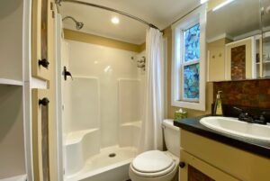 Small bathroom on Just Wandering Lane with a white shower, toilet, sink, mirror, window, and patterned glass.
