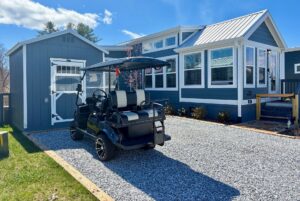 A black golf cart is parked on Mount Meadow Lane in front of a modern blue tiny house with white trim.