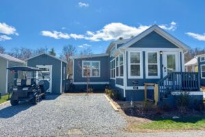 Charming blue tiny house with white trim on Mount Meadow Lane, featuring a porch, shed, and golf cart on gravel driveway.