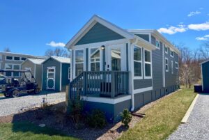 A small gray tiny house with a porch sits on Mount Meadow Lane, framed by clear blue skies and trees.