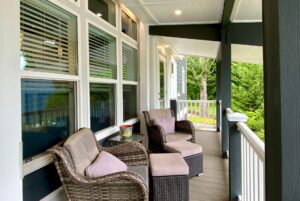 Two wicker chairs with ottomans and cushions on a covered porch at Just Wandering Lane, surrounded by greenery and windows.